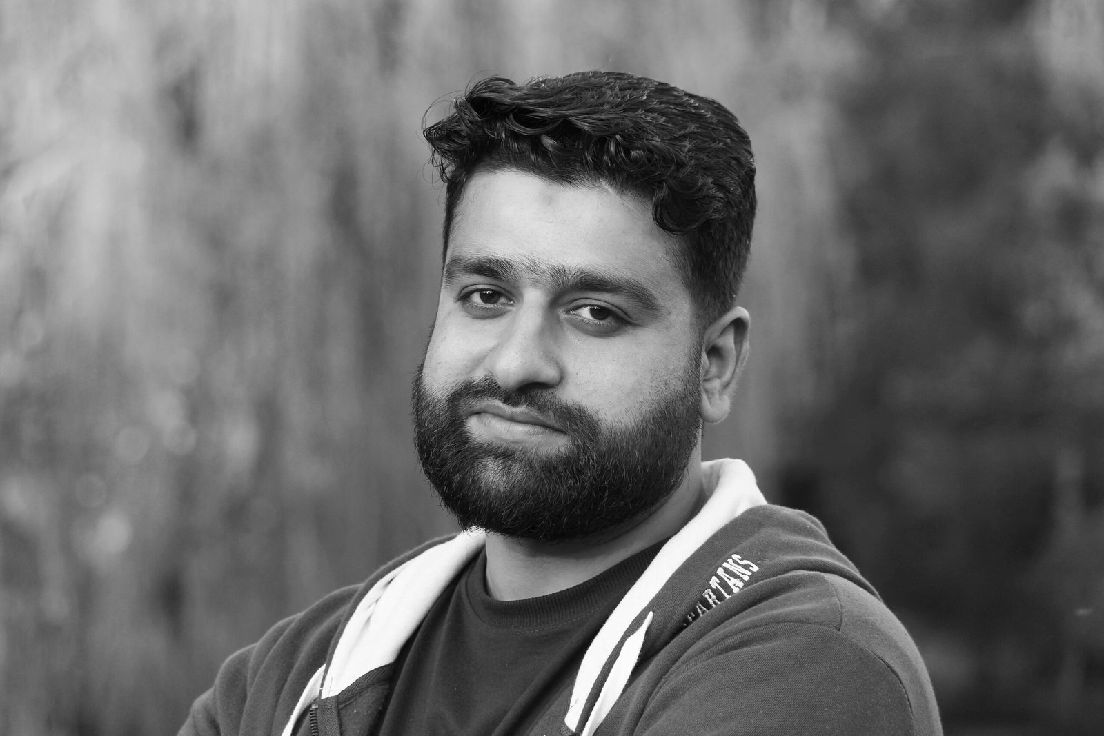 A serious man with a beard in a hoodie poses outdoors. Black and white portrait.